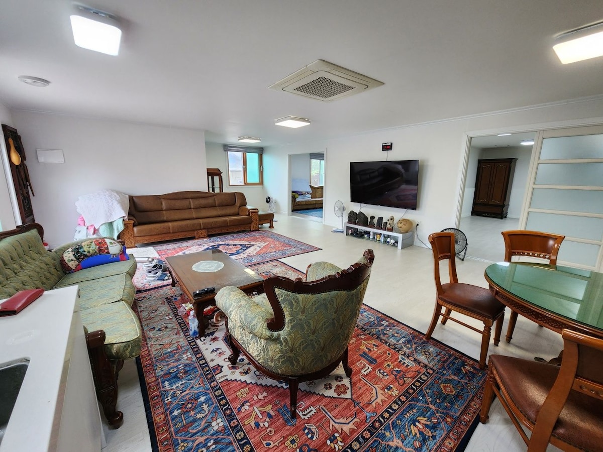 A spacious living area is furnished with a variety of seating options, including a patterned sofa and classic armchairs. A large television is mounted on one wall, and a round dining table is placed nearby. A colorful rug covers the floor, adding warmth to the space.