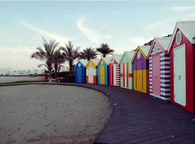 A row of colorful beach huts stands along a sandy shore, each featuring distinct patterns and vibrant hues. Palm trees are visible in the background, and a pathway curves gently alongside the huts, inviting strolls by the beach.