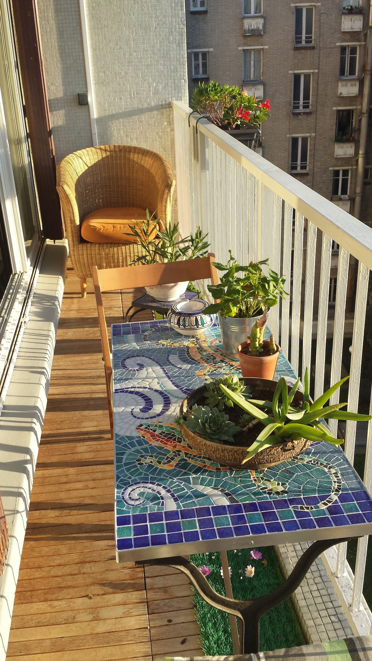 A narrow balcony features a decorated table with colorful mosaic detailing, surrounded by potted plants. A wicker chair is positioned adjacent to the table, creating a space for relaxation. Sunlight highlights the wooden flooring and vibrant greenery, adding to the serene atmosphere.