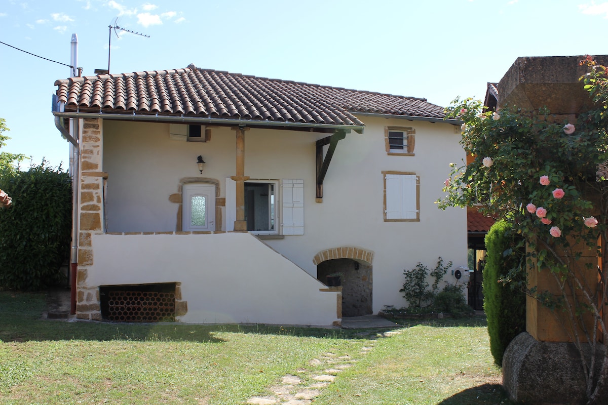 A charming two-story farmhouse is featured, showcasing a tiled roof and whitewashed walls. Ground-level windows are framed with wooden shutters. Outdoor greenery includes a small pathway and blooming roses, enhancing the peaceful setting of the property.