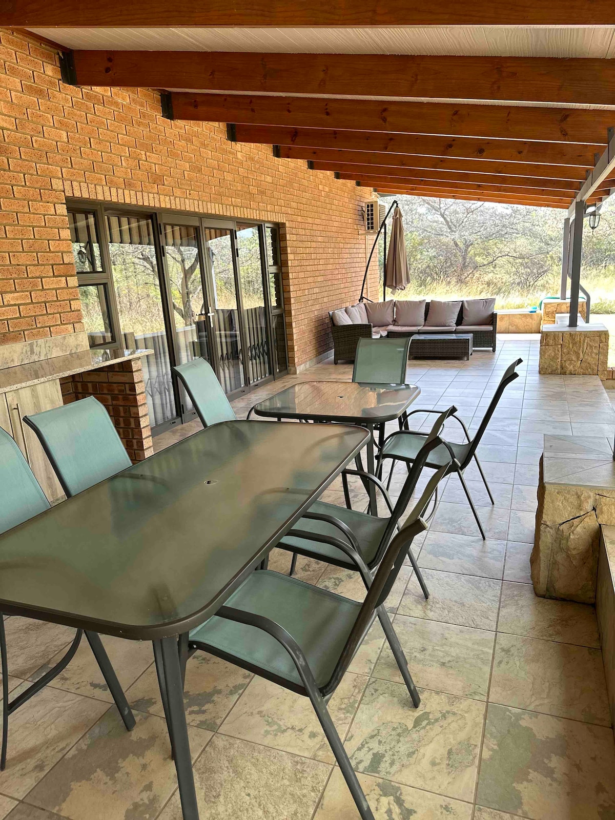 An outdoor patio area features a large table surrounded by several chairs, all set against a backdrop of brick walls and wooden beams. Comfortable seating is positioned nearby, providing a relaxed atmosphere for gatherings. Large glass doors lead into the interior space, allowing natural light.