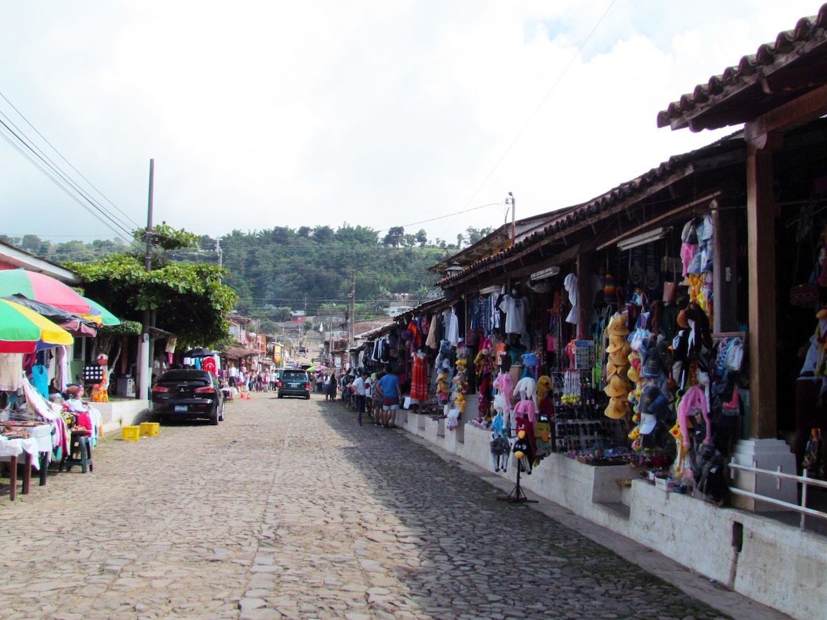 A cobblestone street is lined with stalls offering a variety of colorful clothing and handmade items. Vibrant umbrellas provide shade for shoppers, while local vendors display their goods. Green hills are visible in the background, contributing to the lively atmosphere of the market.