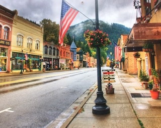 A quiet street is lined with historic buildings, showcasing an array of colors and architectural details. An American flag hangs prominently, while planters with flowers add charm to the sidewalk. Mountains rise in the background, providing a scenic backdrop.