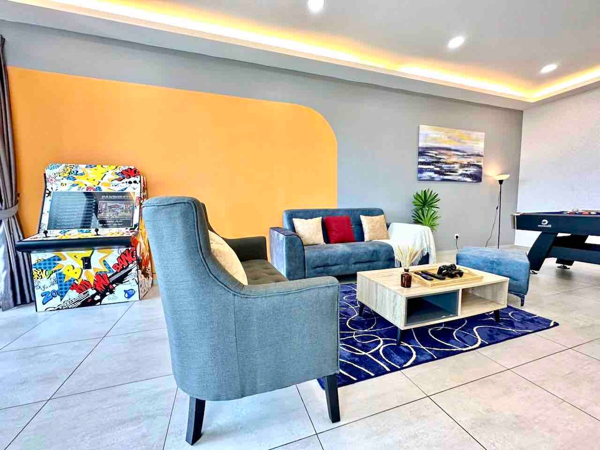 A modern living area features a gray sofa and armchair arranged around a light wood coffee table on a patterned rug. A vibrant orange accent wall complements the space, while an arcade game machine and a foosball table add recreational options.