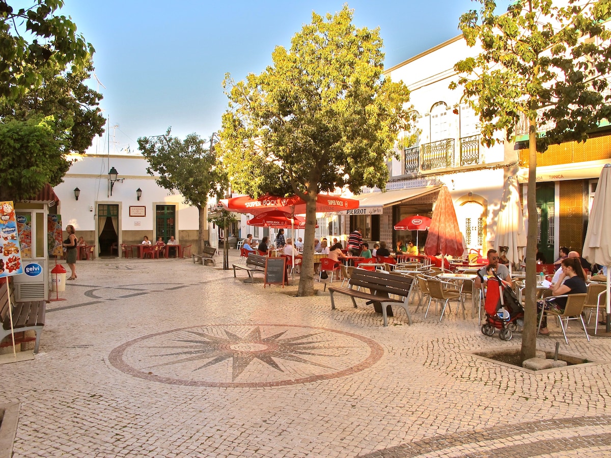 A lively square is set with outdoor seating for cafes under large umbrellas. Paved with decorative tiles, the area features trees providing shade. Patrons are seen enjoying their meals, while the surrounding buildings display traditional architectural details. The atmosphere reflects a vibrant community space.