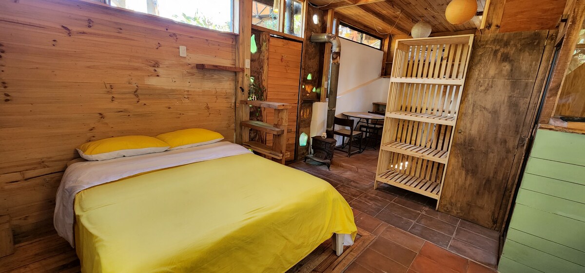 A wooden loft space features a spacious bed with yellow bedding. Nearby, a dining area with a table and chairs is visible. Natural light fills the room through large windows, highlighting the rustic wooden elements and storage shelves.