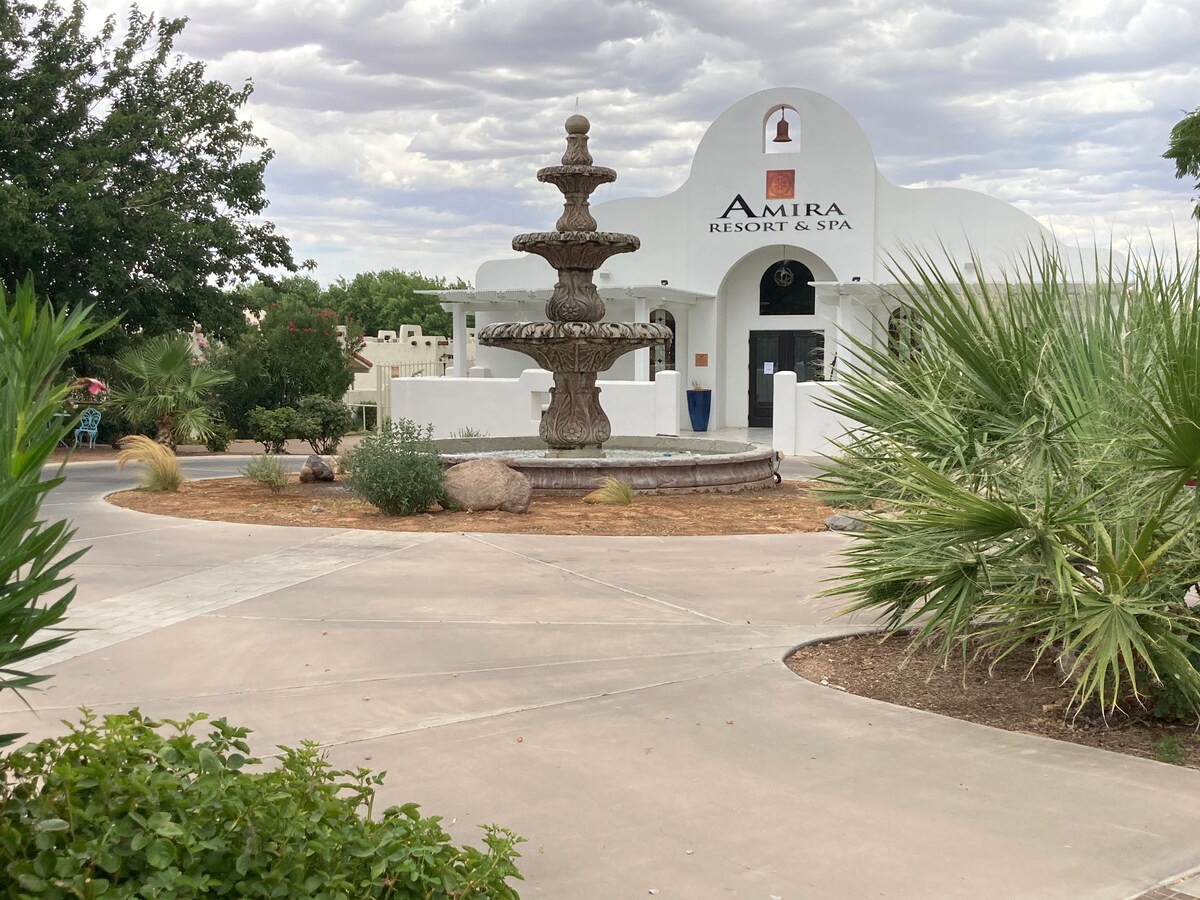 The entrance to Amira Resort & Spa features a decorative fountain surrounded by lush landscaping. Smooth pathways lead to the resort's main entrance, showcasing a white stucco facade with a logo prominently displayed above. Green foliage and rock gardens enhance the serene atmosphere.