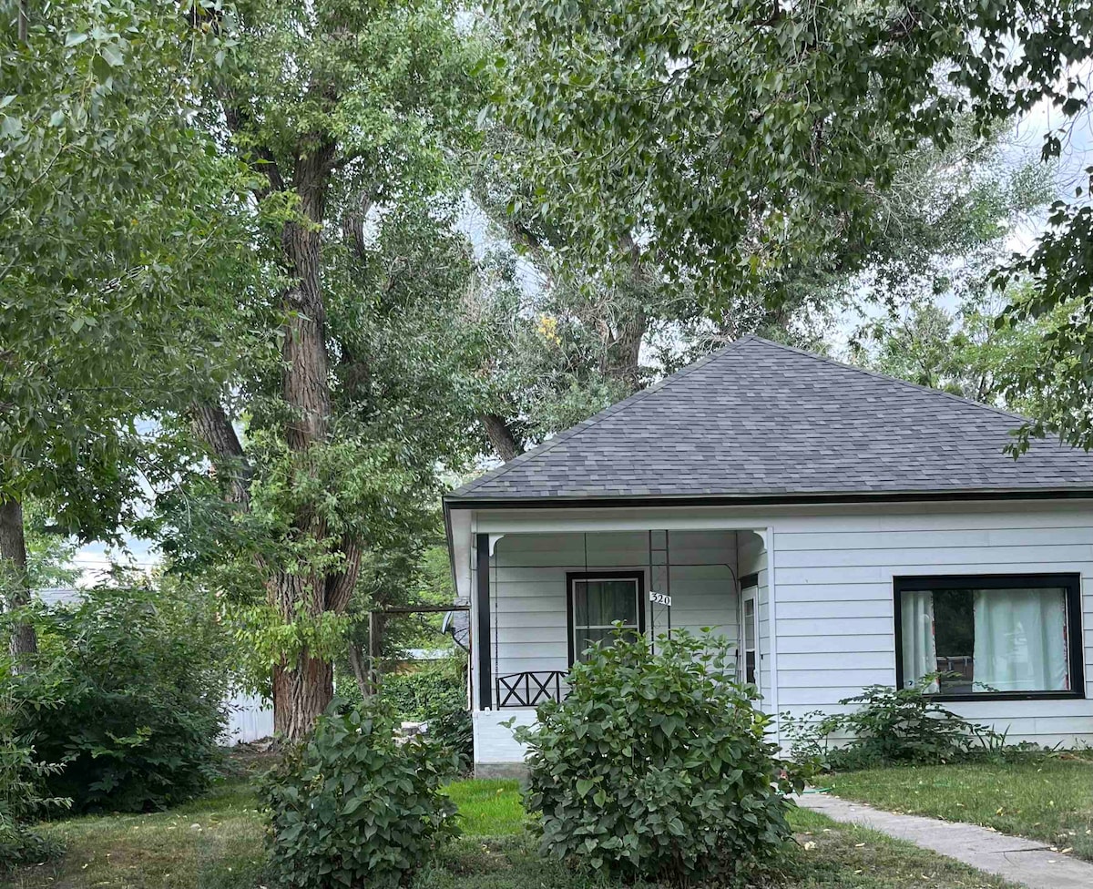 A charming exterior view of a single-story home, featuring a gabled roof and covered porch. Lush greenery surrounds the property, including tall trees and shrubs, creating a tranquil atmosphere. The house's white siding and large windows contribute to its inviting appearance.