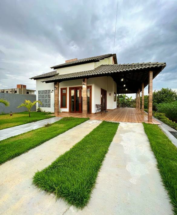 The exterior of the chalé features a welcoming entrance with a covered porch. Lush green grass and smooth concrete pathways lead up to the front doors. The structure is complemented by a traditional roof design, set against a cloudy sky.