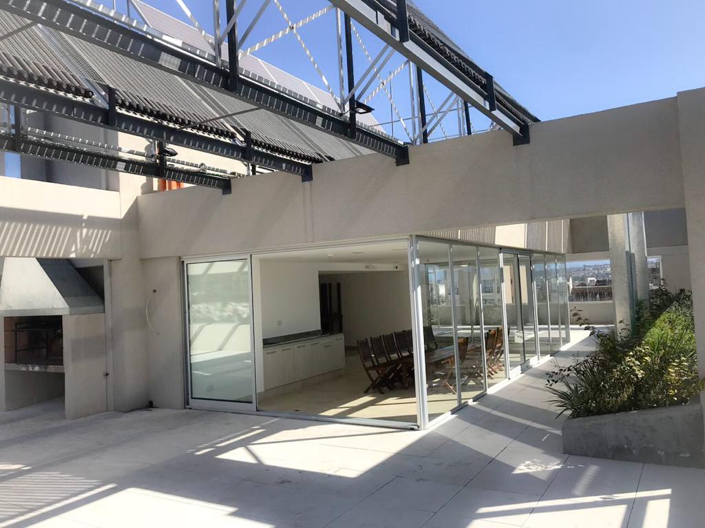 A spacious outdoor terrace is featured, showcasing large glass doors that open to the interior. A dining table with multiple chairs is set up, with a barbecue area visible. Sunlight fills the space, highlighting the modern architectural details and clean lines.