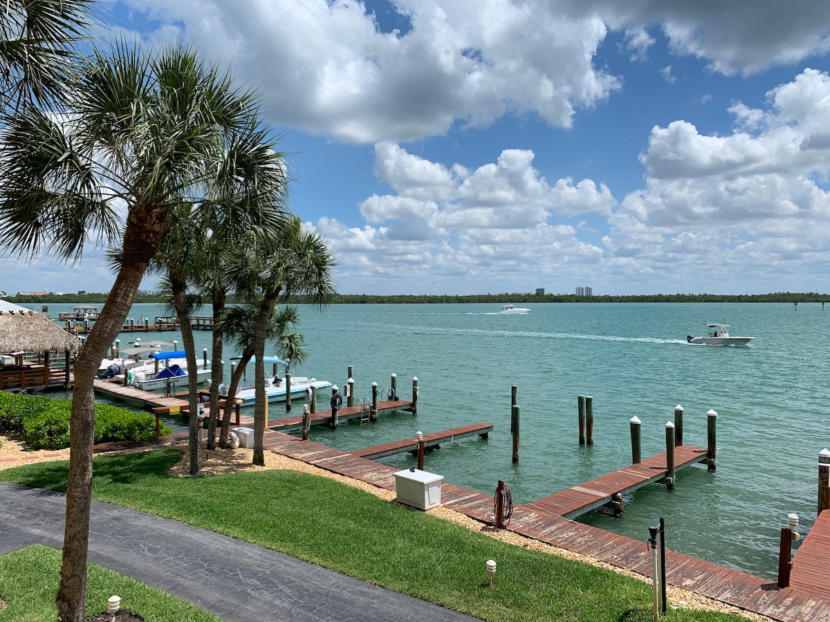A scenic waterfront view features calm turquoise waters and boats moored along wooden docks. Lush palm trees frame the scene, and a paved path leads toward the water's edge. Fluffy clouds drift across the sky, enhancing the tranquil atmosphere of the coastal setting.