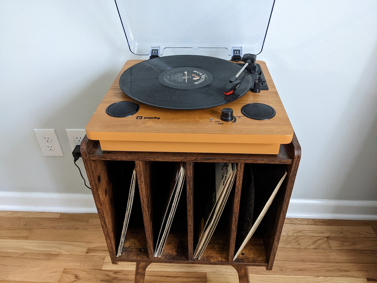 A wooden record player is situated on a mid-century modern storage unit, featuring slots for vinyl records. The player has a clear lid and is styled with a warm wooden finish, while several records are neatly arranged below, showcasing a blend of vintage and contemporary design.