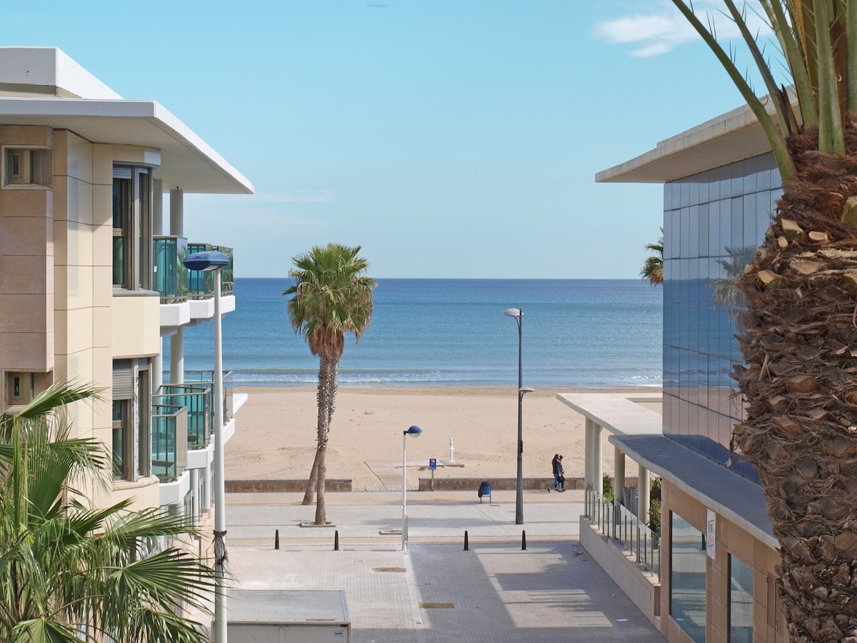 A serene view of the beachfront is captured, showcasing soft sandy shores and calm blue waters. Palm trees line the promenade, while modern buildings frame the scene, creating a sense of coastal tranquility. The sky is bright and clear, inviting relaxation.