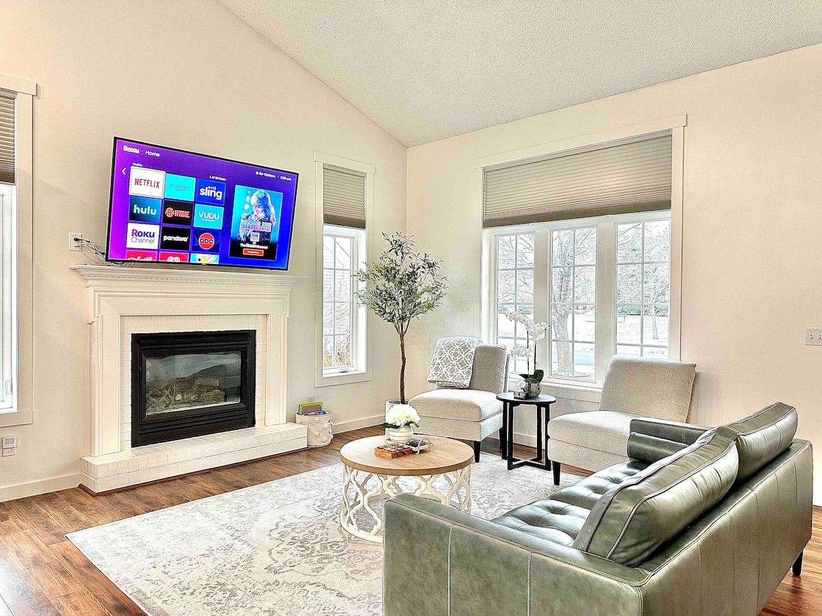 The living room features a cozy seating area with a spacious green couch and two armchairs. A circular coffee table sits in the center. A modern fireplace adds warmth, complemented by a large TV mounted on the wall nearby. Natural light floods through multiple windows.