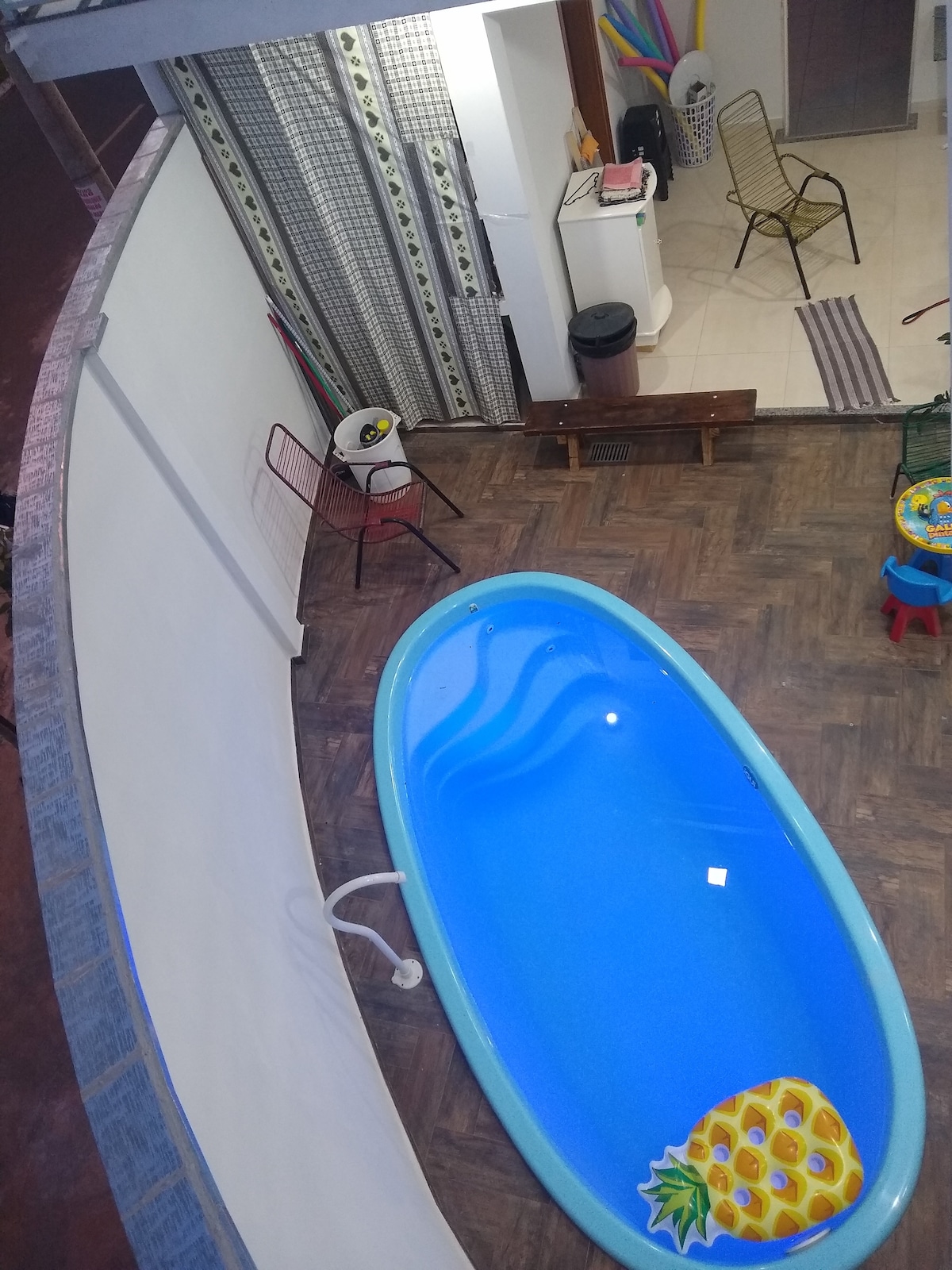 An overhead view of a small indoor pool is visible, featuring a playful pineapple design at one end. Surrounding elements include colorful chairs and a bench, with vibrant decorations seen in the background, contributing to a cheerful atmosphere.