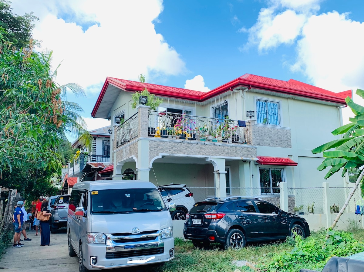 A two-story house with a red roof and a spacious front yard is depicted. Multiple vehicles are parked outside, and lush greenery surrounds the property. A covered porch with decorative railings adds charm to the entrance, highlighted by a clear blue sky above.