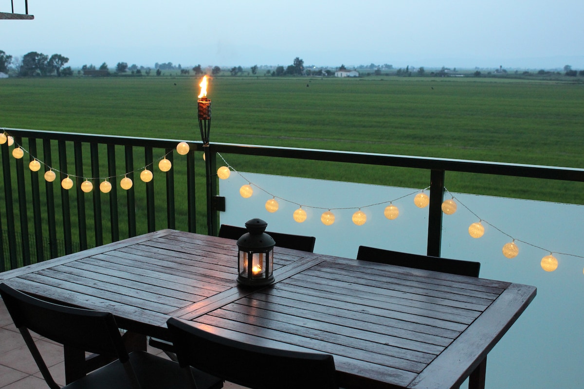 A balcony dining area is presented with a wooden table and chairs, illuminated by a lantern and soft string lights. A scenic view of green fields is visible beyond the railing, creating a serene ambiance for outdoor dining.