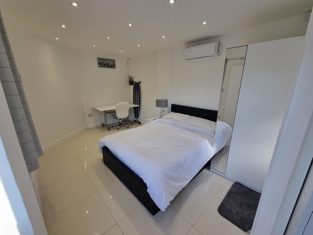 A bright sleeping space features a spacious bed covered with a white duvet. A desk and chair are positioned nearby, and a full-length mirror stands against the wall. Recessed lighting provides illumination, while a soft rug lies on the glossy tiled floor.