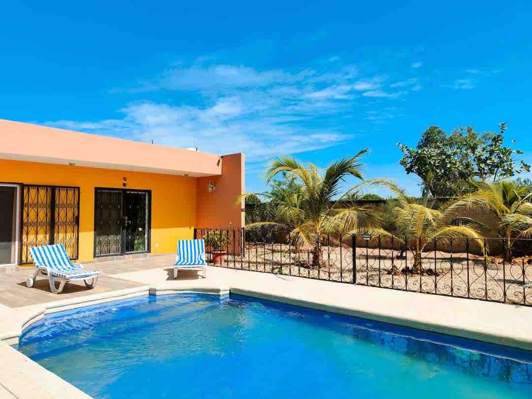 A private pool is surrounded by a sun deck featuring two lounge chairs. The vibrant orange exterior of the villa offers a contrast to the clear blue sky. Tropical greenery and palm trees create a relaxed outdoor setting.
