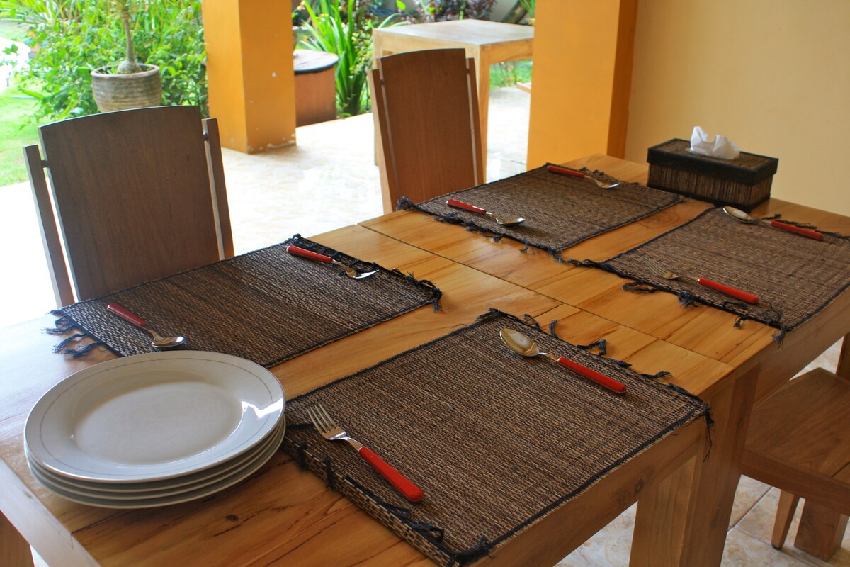 A dining table is set with wooden chairs, featuring woven placemats. Each setting includes a plate, knife, and fork with red handles. A small box for tissues is visible on the table. Outdoor greenery can be seen in the background, enhancing the dining environment.