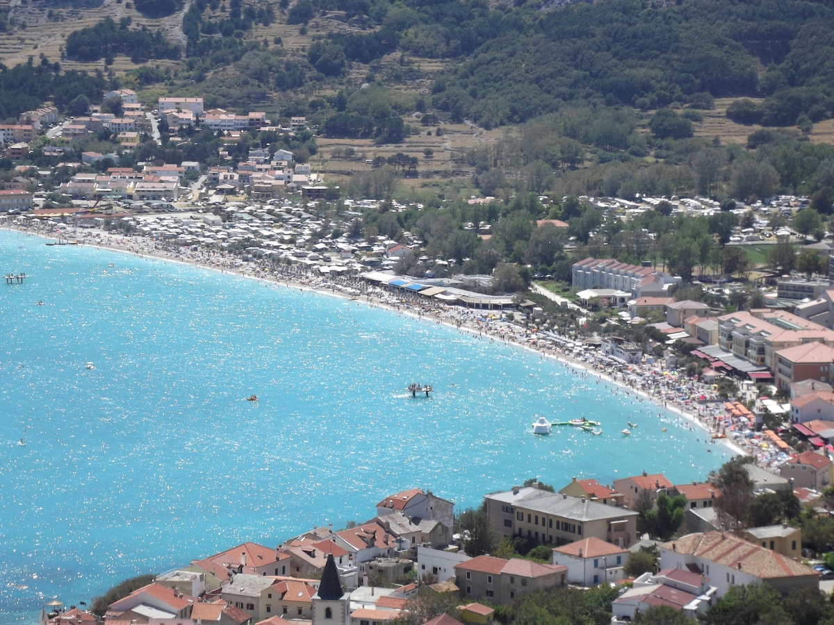 A panoramic view of a vibrant beach reveals a long stretch of sandy shore, with numerous sunbathers and beachgoers. The clear turquoise water reflects sunlight, while surrounding hills and greenery frame the scene. A cluster of beach umbrellas and boats can be seen dotting the water.