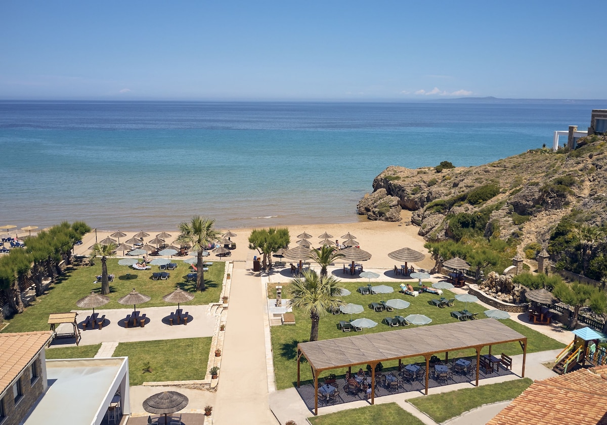 A sandy beach stretches along the shoreline, complemented by a clear blue sea. Palm trees and shaded lounge areas are scattered across the grassy section leading to the beach. A wooden pergola provides additional seating with views of the landscape.