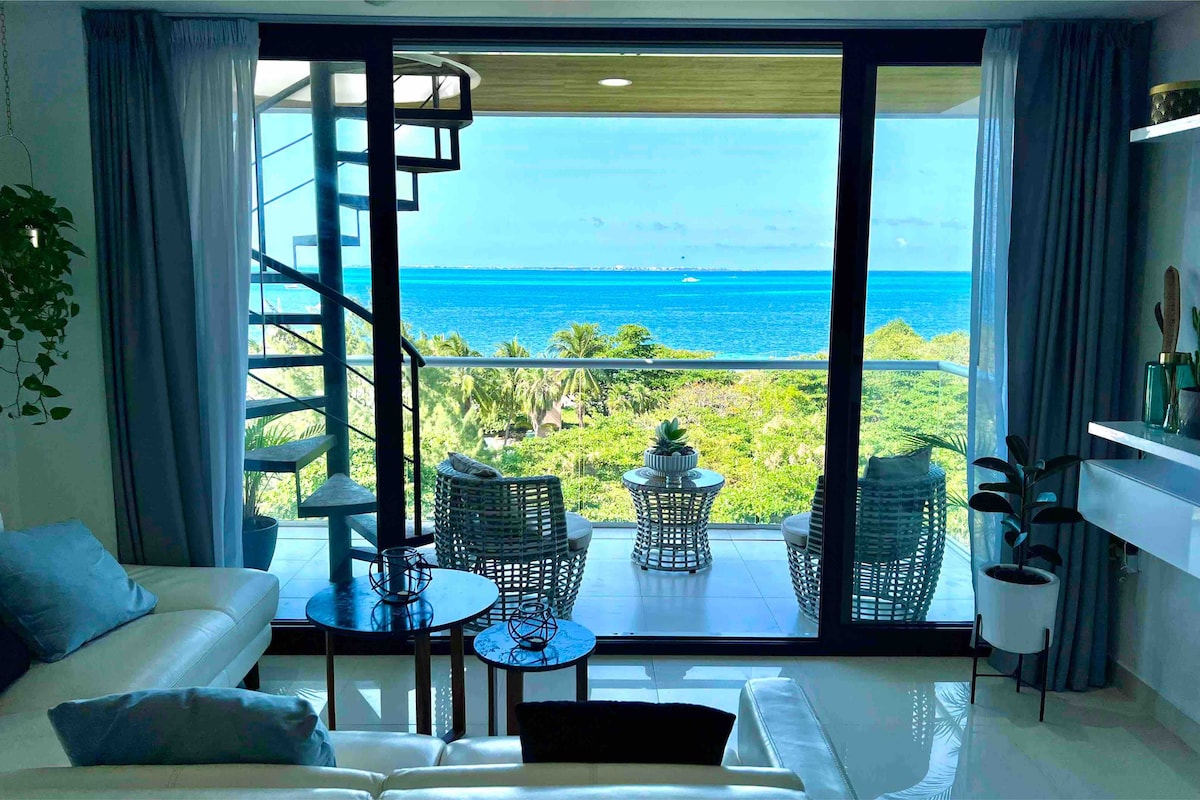 A modern living space features a spiral staircase leading to a balcony with stunning ocean views. Large glass doors provide natural light, while a comfortable white couch and side tables create a relaxing atmosphere. Lush greenery is visible outside, complementing the blue waters.
