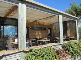 A covered outdoor dining area features wooden beams and a shaded roof. Tables and chairs are arranged under the shelter, with greenery visible around the perimeter. A sign displaying 'Bar' adds a welcoming touch, while large windows invite natural light into the space.