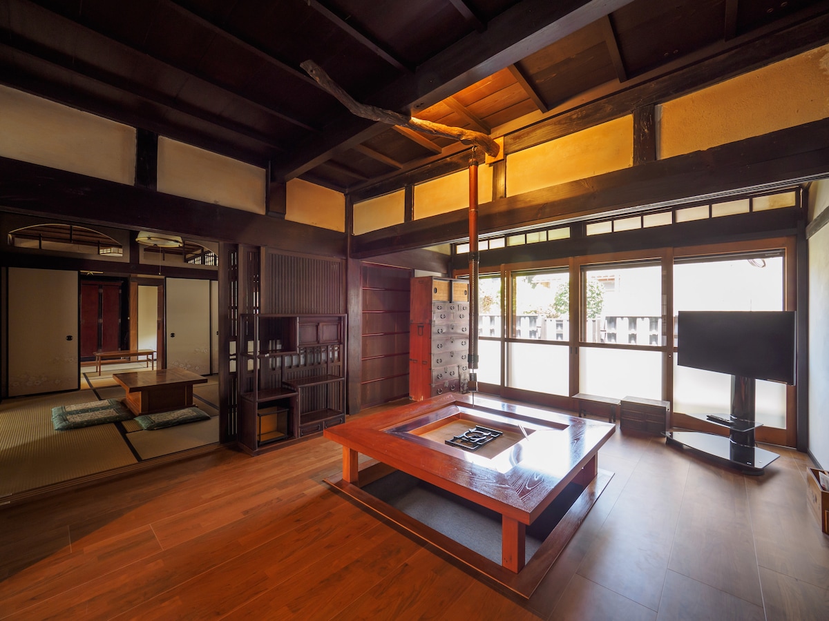 A spacious living area is highlighted by a low wooden table placed over polished flooring. Traditional wooden beams support the ceiling, while large windows allow natural light to flow into the space. A television is positioned in the corner, complementing the cozy ambiance.