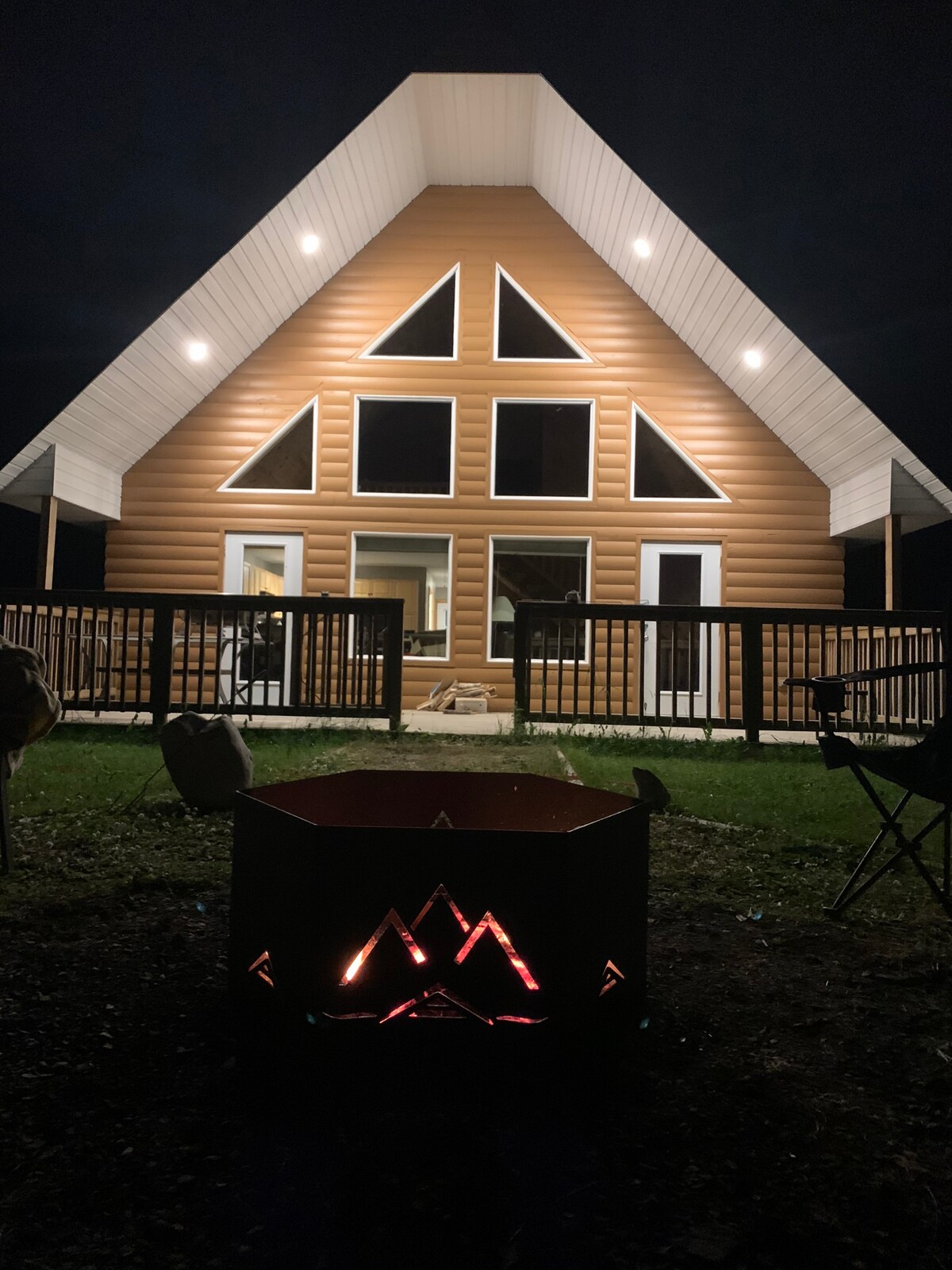 A modern A-frame cabin is illuminated at night, showcasing large triangular windows that reflect the soft glow of outdoor lighting. A fire pit, with geometric designs, captures the warmth of flames in the foreground, inviting relaxation and gathering.
