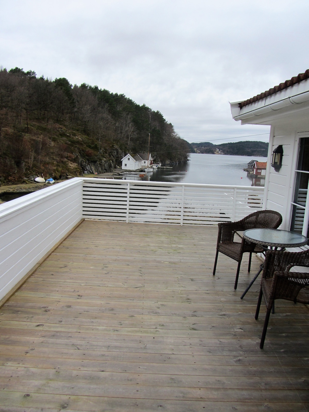 A wooden deck features a small round table and two wicker chairs, overlooking serene water surrounded by green hills. The white railing provides a clear view of the calm bay, contributing to a peaceful outdoor setting.