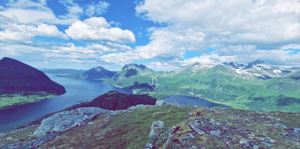 A panoramic view captures mountains and a serene body of water surrounded by lush greenery. The sky is dotted with clouds, and sunlight bathes the landscape, revealing the natural beauty of the Helgelandskysten area.