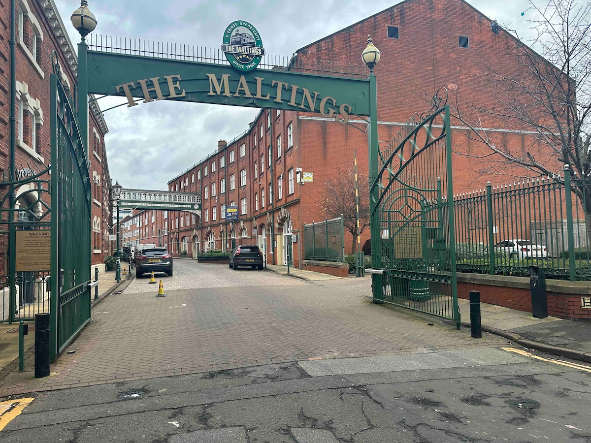 An elegant entrance to The Maltings is showcased, featuring a grand archway with the name prominently displayed. The paved pathway leads into a gated area, flanked by brick buildings on either side and trees lining the ground, creating an inviting approach.