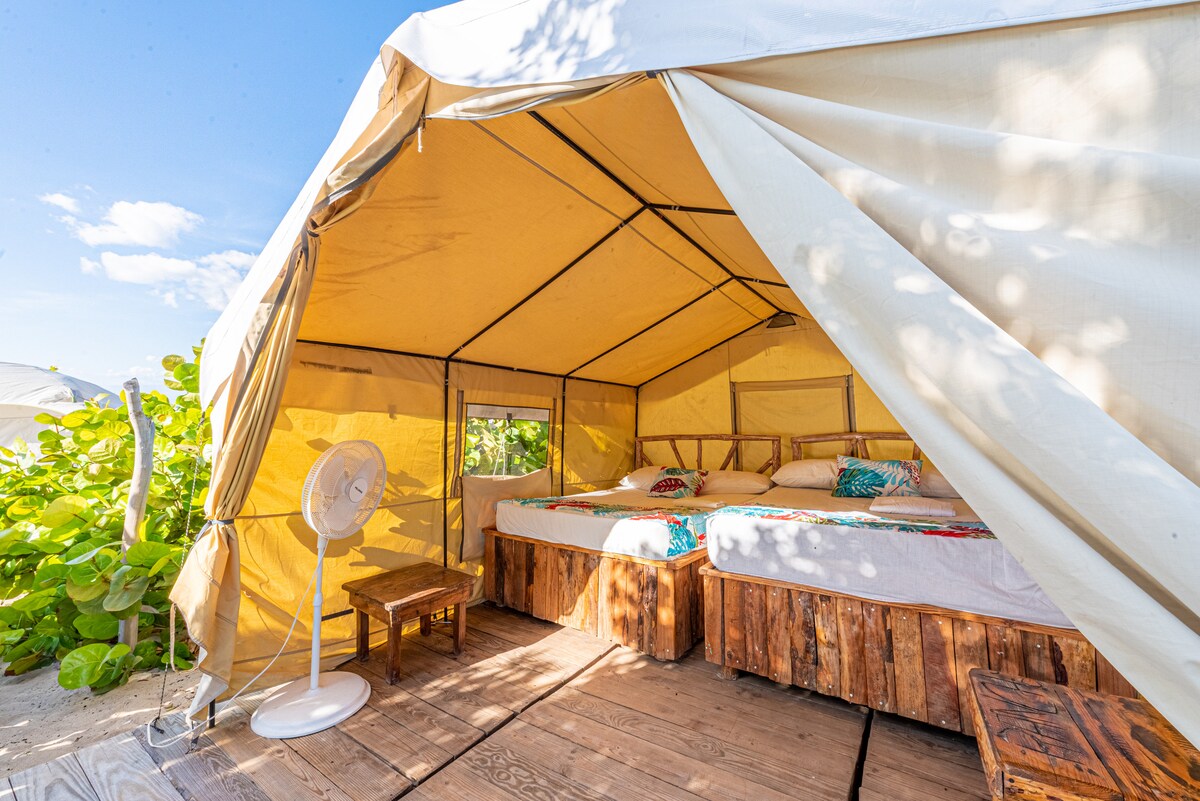 A glamping tent features two spacious beds positioned on a wooden platform, surrounded by natural greenery. A simple fan provides cooling comfort, and colorful textiles add a touch of detail. The light filters through the open entrance, creating a bright and inviting space.