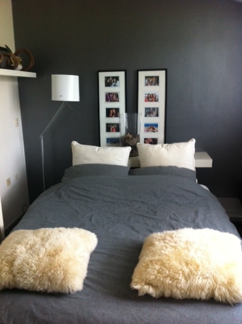 A cozy sleeping area features a spacious bed dressed in gray bedding, flanked by two soft pillows. The wall is painted in a dark tone, and framed photographs add a personal touch. A minimalist lamp stands nearby, completing the warm, inviting space.