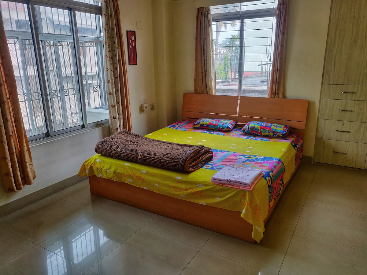 A spacious bedroom features large windows allowing natural light to fill the room. A comfortable double bed is adorned with colorful bedding and soft pillows. A cozy throw is neatly placed on the bed, and a storage unit complements the light walls.