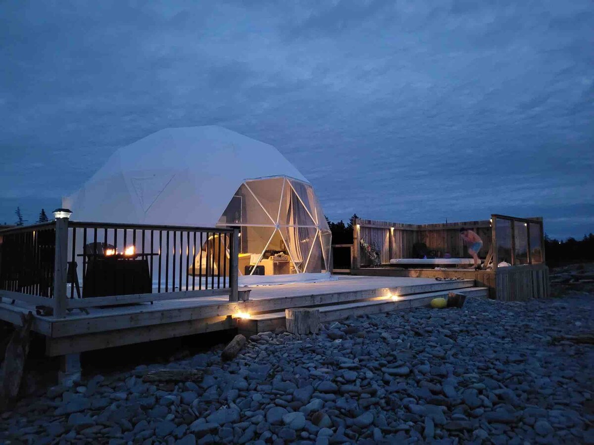 A geodesic dome structure is situated on a rocky beach, illuminated by soft outdoor lighting. The deck features a wooden railing and offers space for relaxation. A jetted hot tub is visible on the deck, surrounded by natural stone, creating a serene coastal atmosphere.