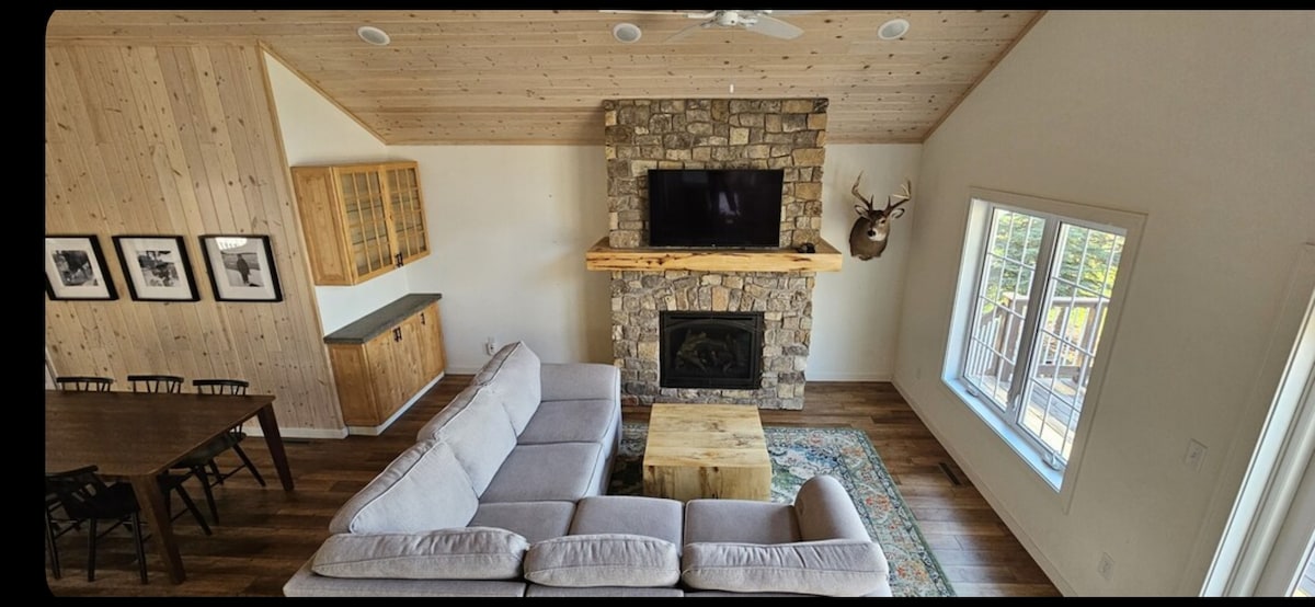 A spacious living area features a large sectional sofa facing a stone fireplace beneath a mounted television. Natural light filters through a nearby window, illuminating a wooden table with seating. Decorative framed artwork is displayed on the walls, enhancing the rustic ambiance.