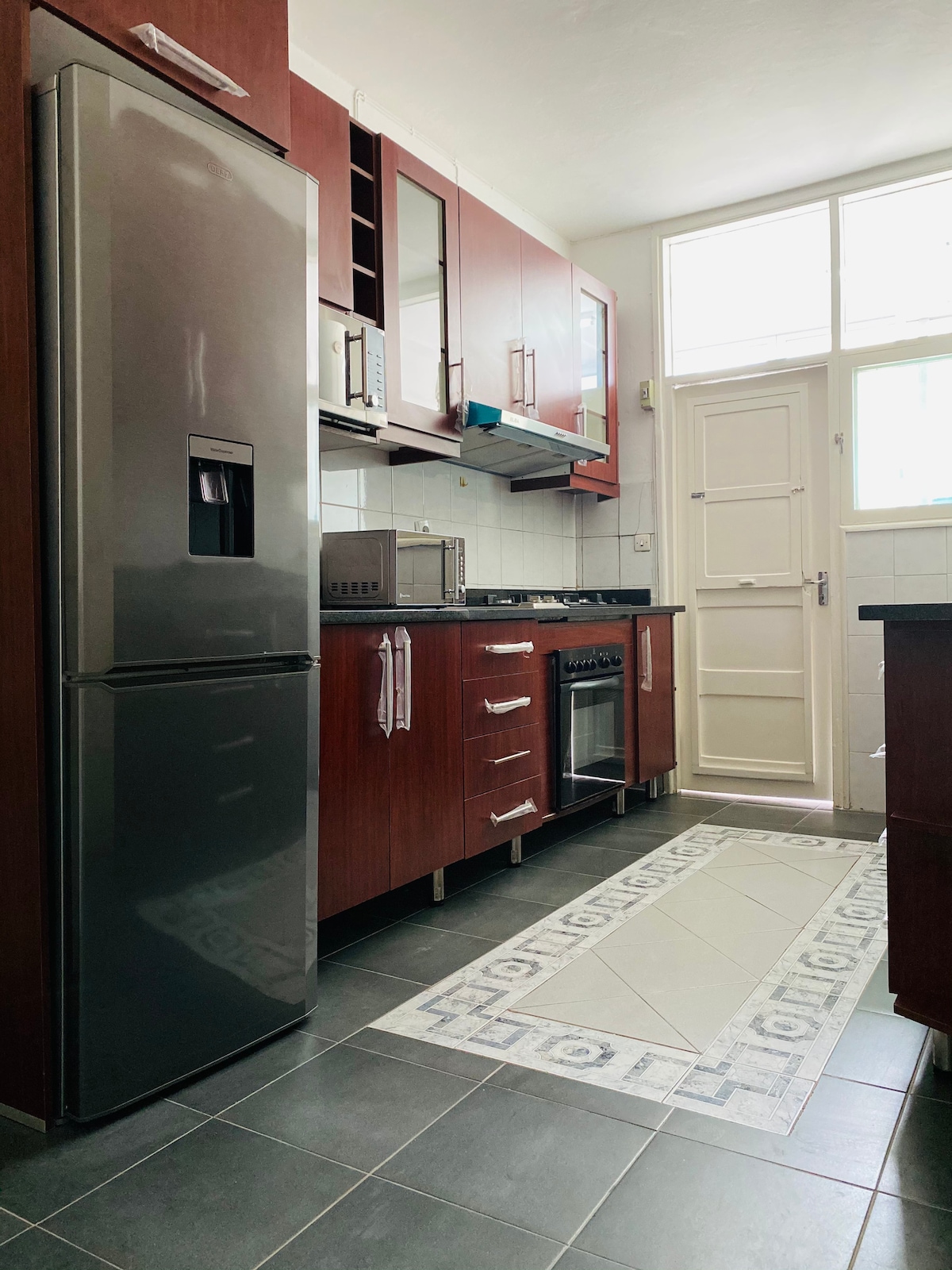 The kitchen features modern wooden cabinetry and stainless steel appliances, including a refrigerator and stove. Natural light enters through multiple windows, illuminating the space. A patterned rug adds a touch of design to the tiled floor, enhancing the practical layout of the room.