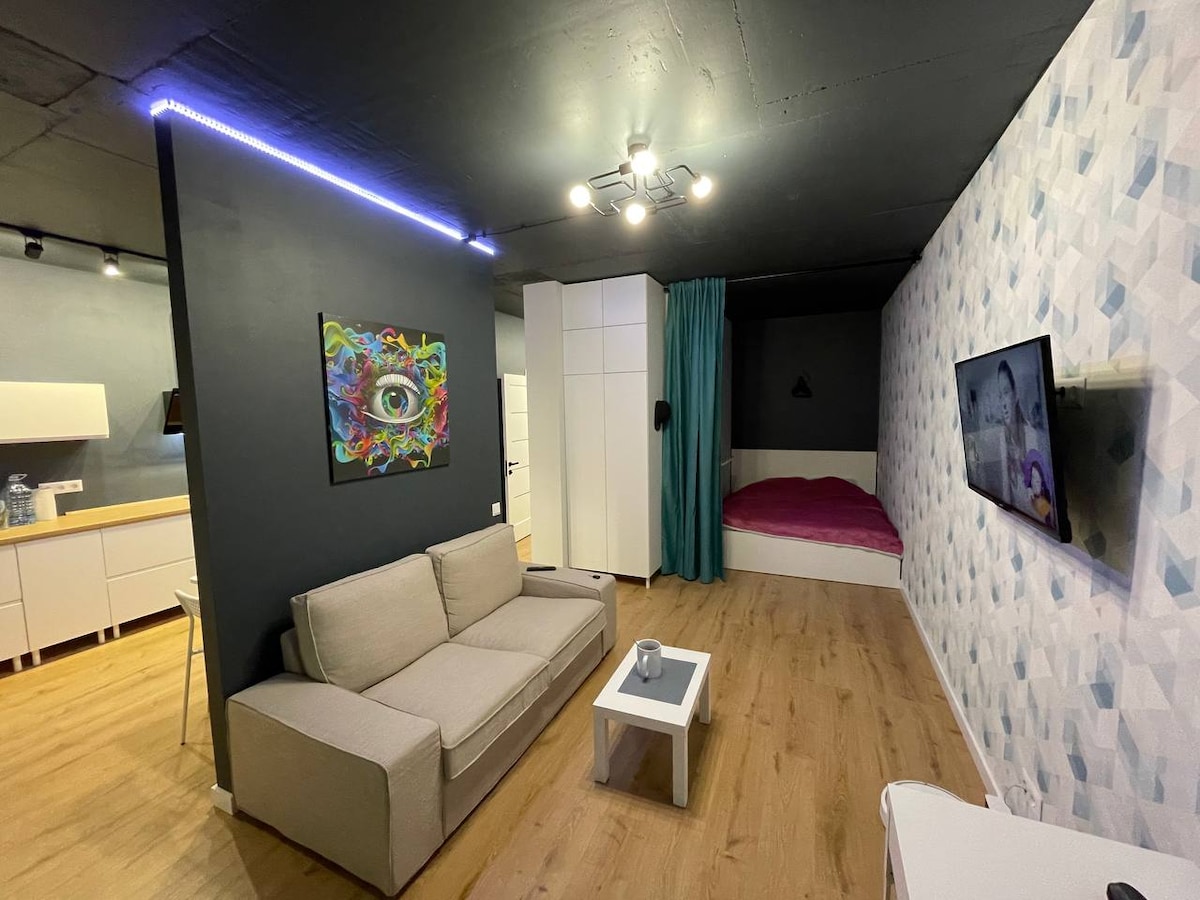 A modern open-plan living space features a light-colored sofa adjacent to a small white table. A colorful wall art piece draws attention on a dark wall. A sleeping area with a spacious bed is partially visible, separated by a green curtain. Soft lighting illuminates the room.