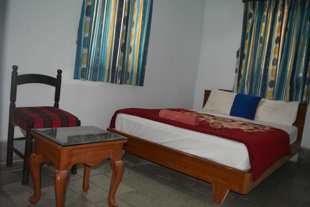 A simple sleeping area features a bedsheet with subtle patterns, a plush red throw, and a colorful accent pillow. A chair with a patterned seat and a glass-topped wooden table are positioned nearby, with light filtering through striped curtains covering the window.