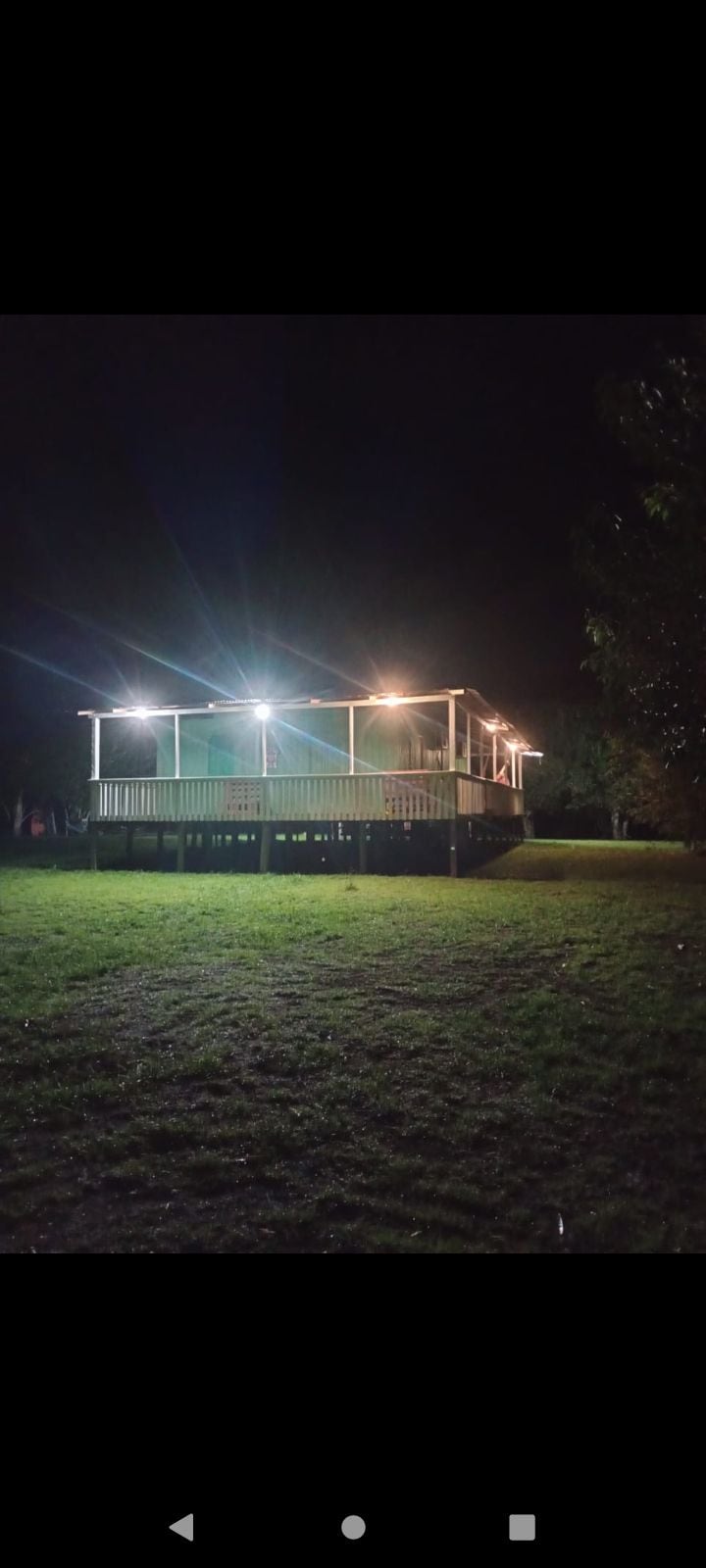 A two-story house, elevated above the ground, is illuminated under the night sky with soft lights. The surrounding grassy area is visible, enhancing the tranquil outdoor setting. The structure features a spacious deck, providing a welcoming area for relaxation.
