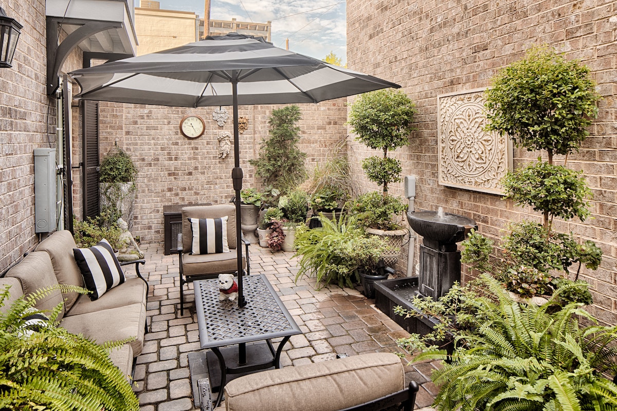 A private courtyard is surrounded by lush greenery and stone walls. Comfortable seating is provided beneath a large umbrella, with a decorative wall feature visible. A tranquil fountain enhances the serene atmosphere, and cobblestone flooring creates a cozy, inviting space.