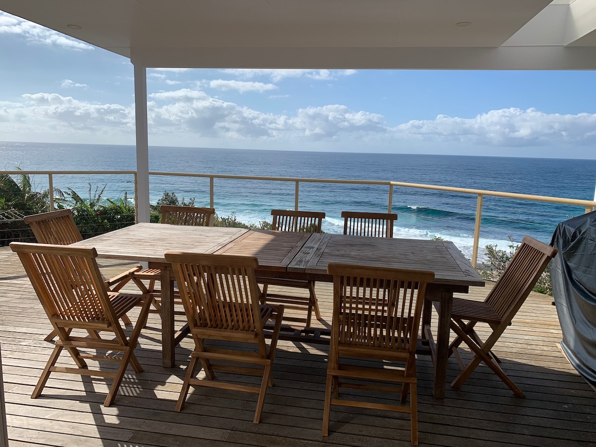 A wooden outdoor dining set is positioned on a deck overlooking the ocean. The table accommodates six chairs with a scenic view of the coastline and gentle waves. The space is bathed in natural light, complemented by a bright blue sky with scattered clouds.
