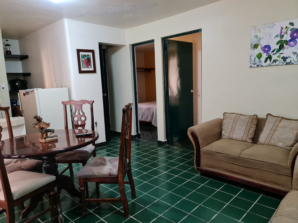 The living space features a round dining table with four chairs and a cozy sofa. Green tile flooring contrasts with the light-colored walls. Two doors lead into private areas, and a small framed artwork adds a touch of color to the room.