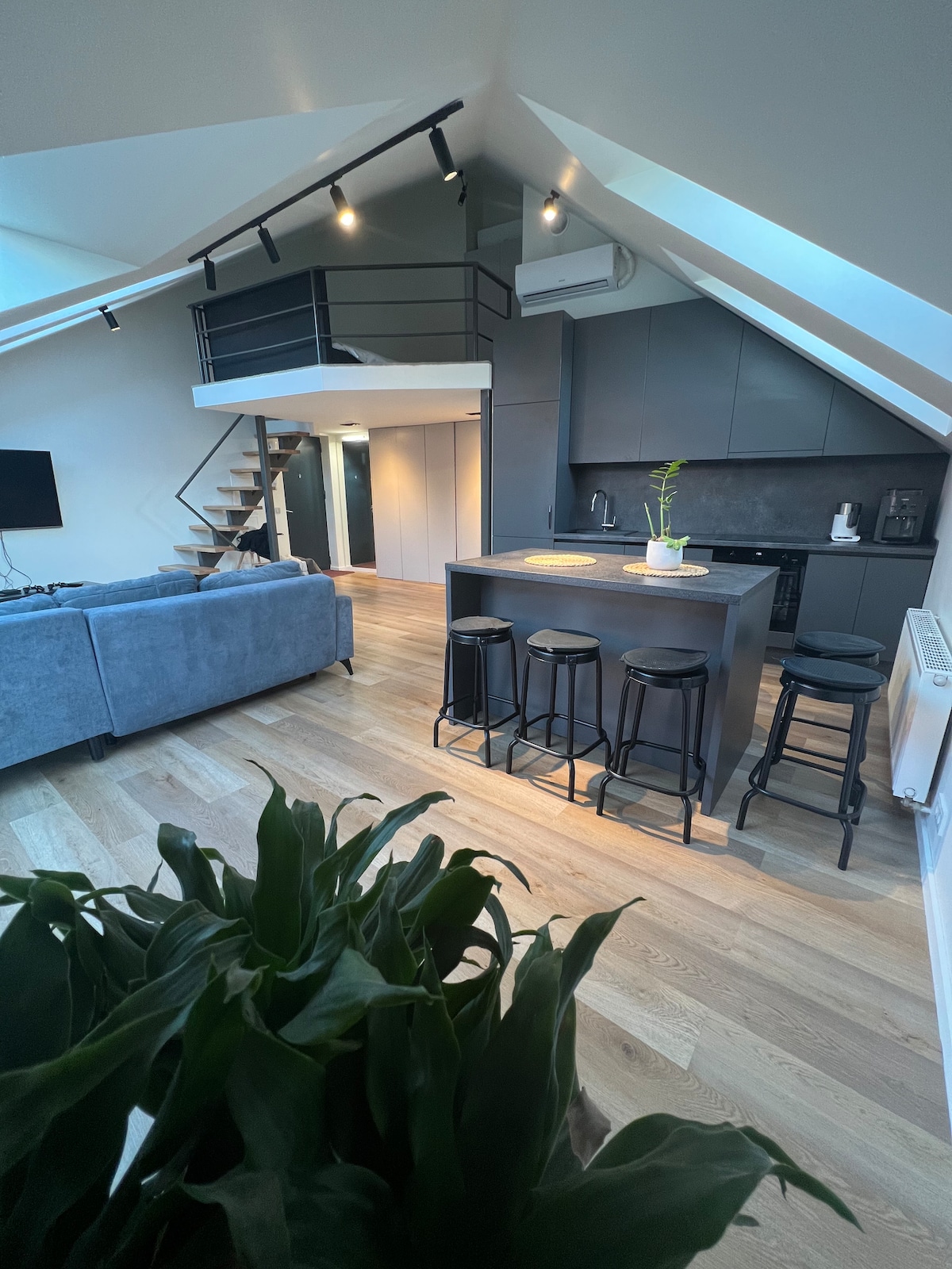 An open-plan loft features a modern kitchen with a central island and four high stools. A cozy seating area is visible, and a staircase leads to a lofted space above. Natural light floods the room through angled skylights, highlighting the sleek cabinetry and wooden flooring.