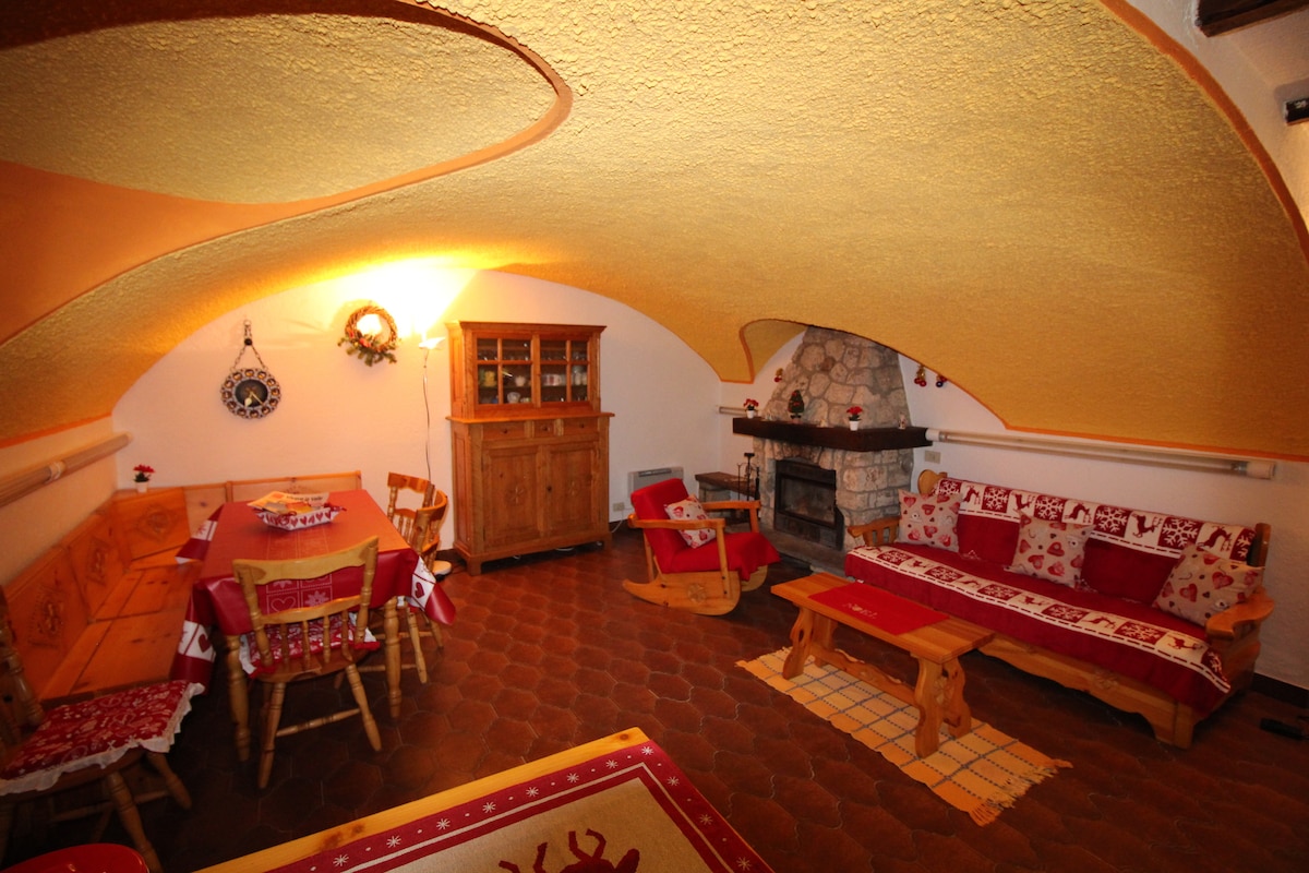 A warm, rustic living area is characterized by a curved golden ceiling and stone fireplace. Wooden furniture, including a dining table and multiple seating arrangements, is adorned with traditional red and white textiles. Natural light is gently diffused throughout the space.