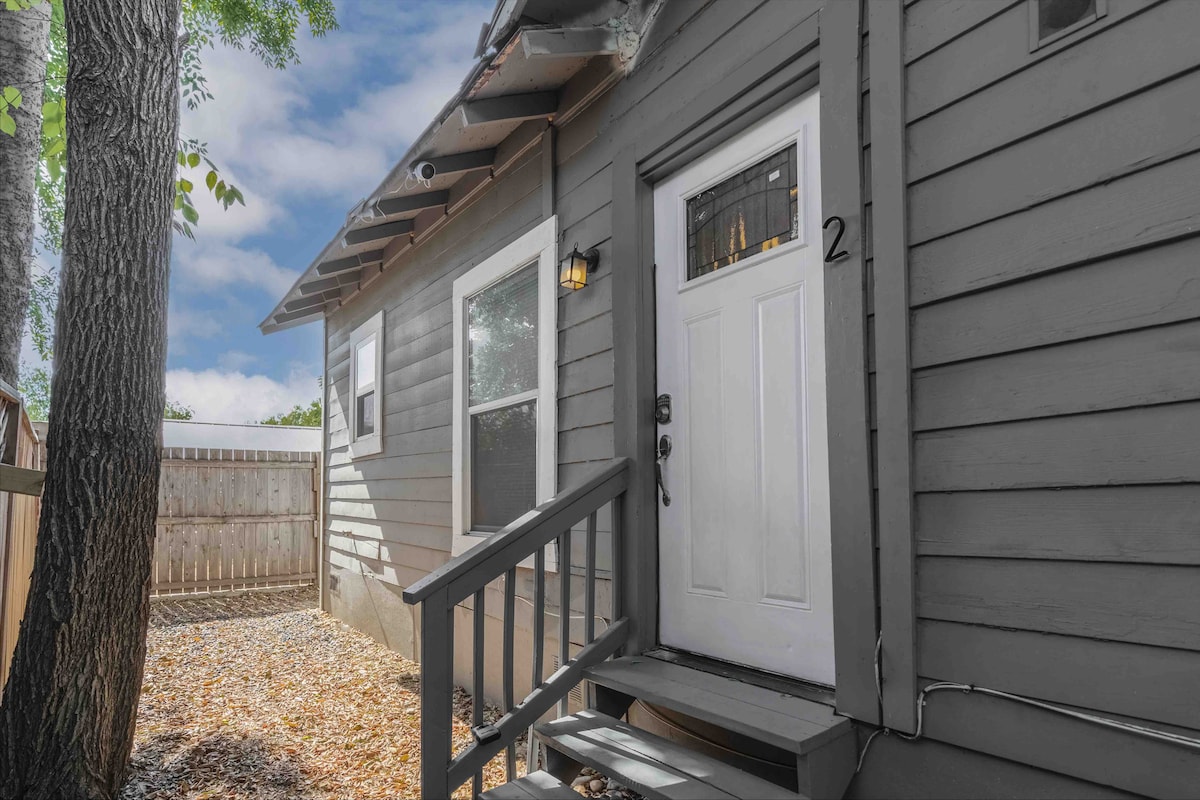 The entrance to unit #2 is highlighted by a welcoming white door, framed by a modest porch and steps. Soft light is provided by a small outdoor fixture. Surrounding greenery and gravel offer a peaceful setting in a quiet neighborhood.
