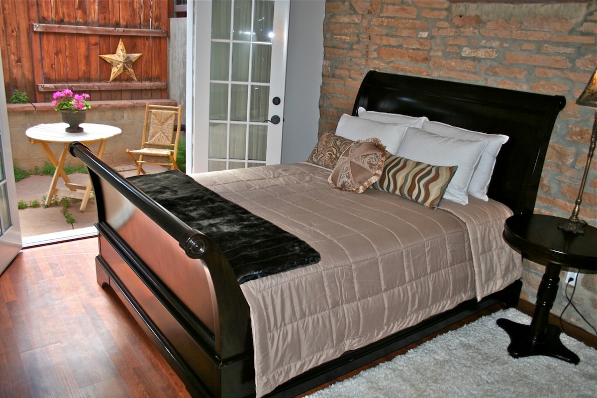 A bedroom features a dark wood sleigh bed adorned with neutral-toned linens. Natural light enters through a glass door leading to a private patio, where a small round table and a chair are visible. Exposed brick walls add character to the room.