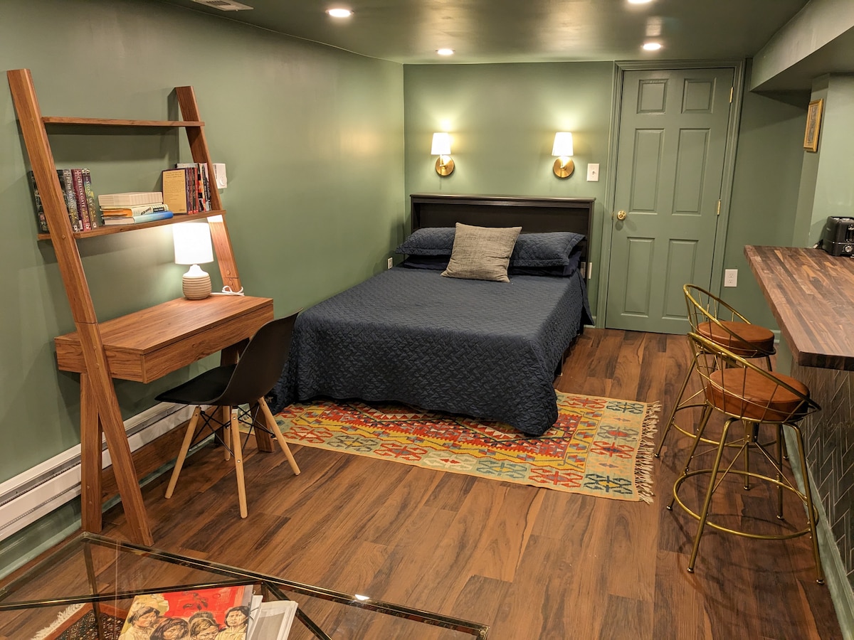 A spacious studio apartment is highlighted by a comfortable full-size bed against a green wall. A wooden desk and sleek chair are positioned beside the bed, with a small lamp providing warm light. A bar area with two stools offers additional seating options.